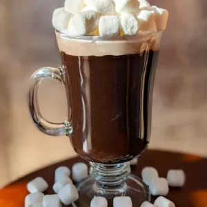 A clear glass mug filled with rich, dark hot chocolate, topped with a generous amount of mini marshmallows, placed on a wooden surface with additional marshmallows scattered around, against a softly blurred background.